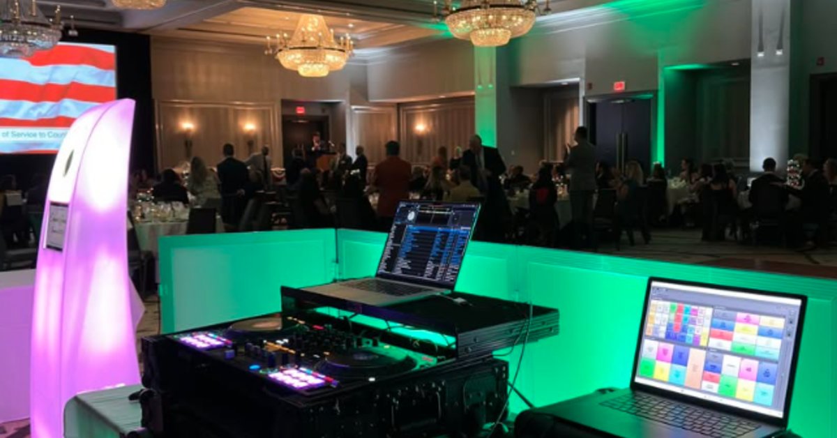 A DJ booth setup at a formal banquet event with glowing green lighting, laptops and audio equipment in the foreground, and guests seated at round tables in a ballroom.