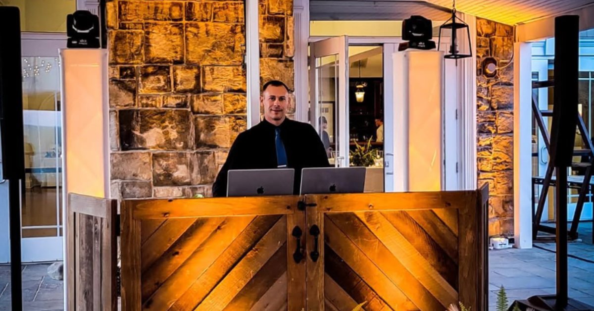A DJ stands behind a rustic wooden booth with laptops and speakers set up at an event space featuring stone walls and warm lighting.