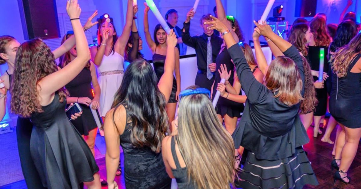 A crowded dance floor at a party with teenagers dancing and raising illuminated foam sticks under colorful club-style lighting.