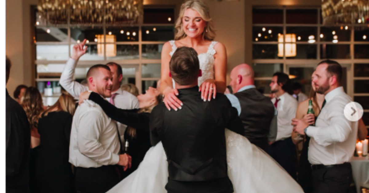 A bride in a white dress smiling while the groom lifts her on a crowded indoor wedding dance floor during a reception.