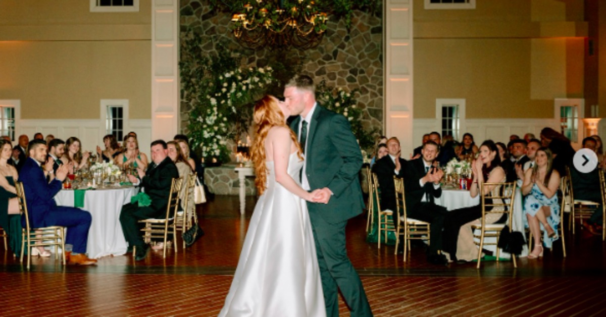 A couple in formal wedding attire holding hands while dancing on a wooden floor as seated guests watch.