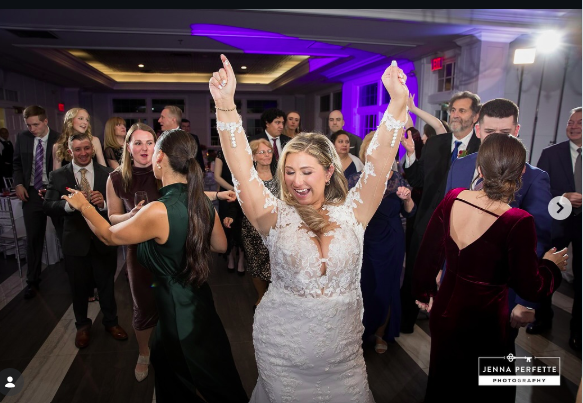 A bride in a lace wedding dress dancing with raised arms among guests under purple lights in a reception hall.