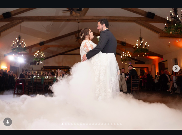 A couple in wedding attire dancing together above low white fog in a warmly lit reception room with chandeliers.